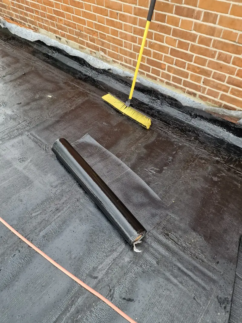 Torch-on felt being laid on a flat roof against a brick wall in Witham, with a roll of cap sheet ready