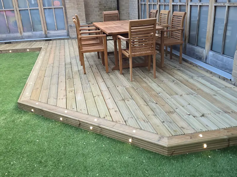 Angled timber deck with inset LED lights, edged by artificial grass beside tall arched windows in Colchester