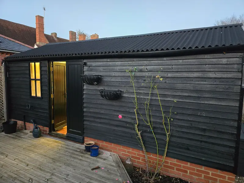 Black timber-clad garden building with a corrugated roof, brick plinth and lit glazed door in a Coggeshall garden