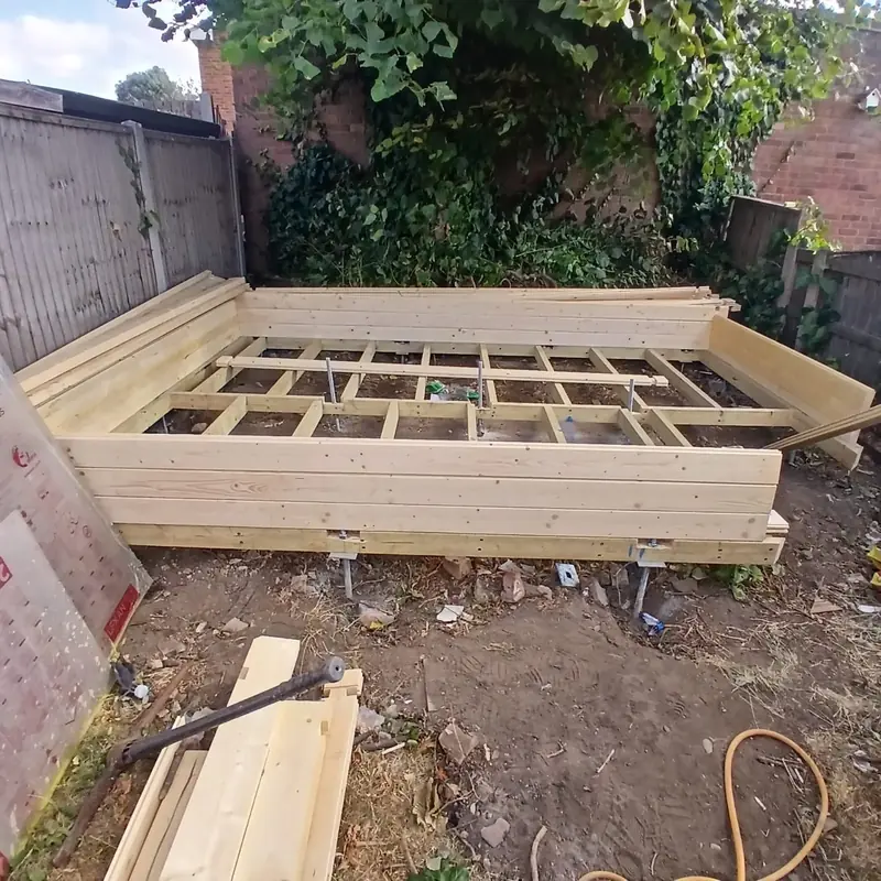 Timber floor frame of joists raised on adjustable galvanised steel feet in a garden corner, wall logs stacked on its edges