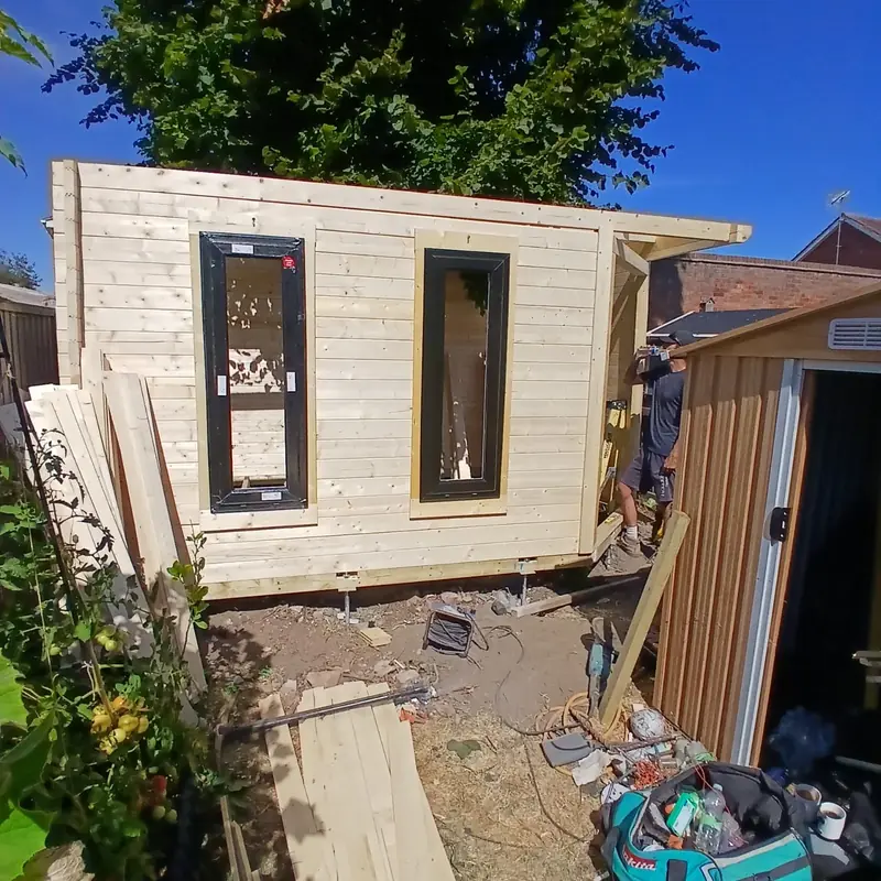 Log garden office walls on adjustable steel feet with two tall anthracite grey windows fitted and the roof beams overhanging