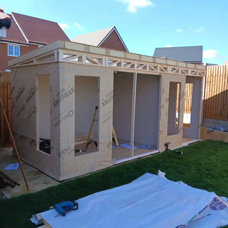 Garden office frame wrapped in breather membrane, with roof joists and insulation fitted, beside brick houses