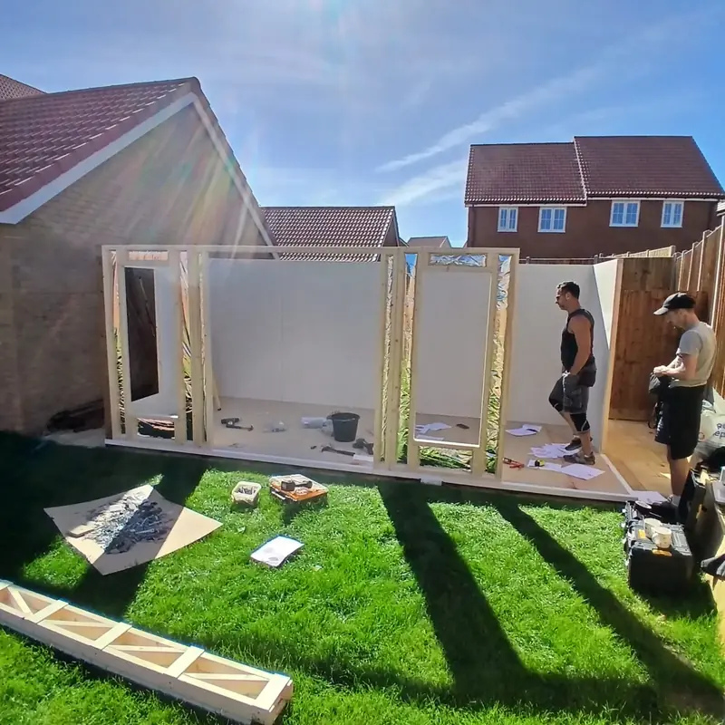 Two installers inside a timber stud frame lined with plasterboard and foil insulation, on a lawn beside new-build houses.