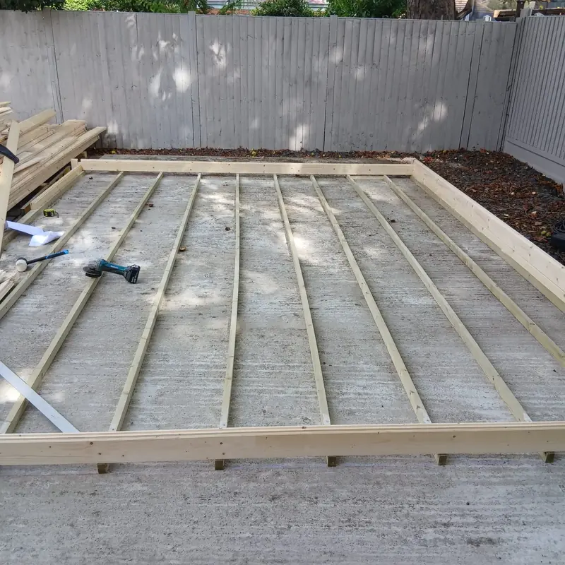 Timber floor bearers spaced evenly across a concrete slab inside the first course of logs, with a grey fence behind.