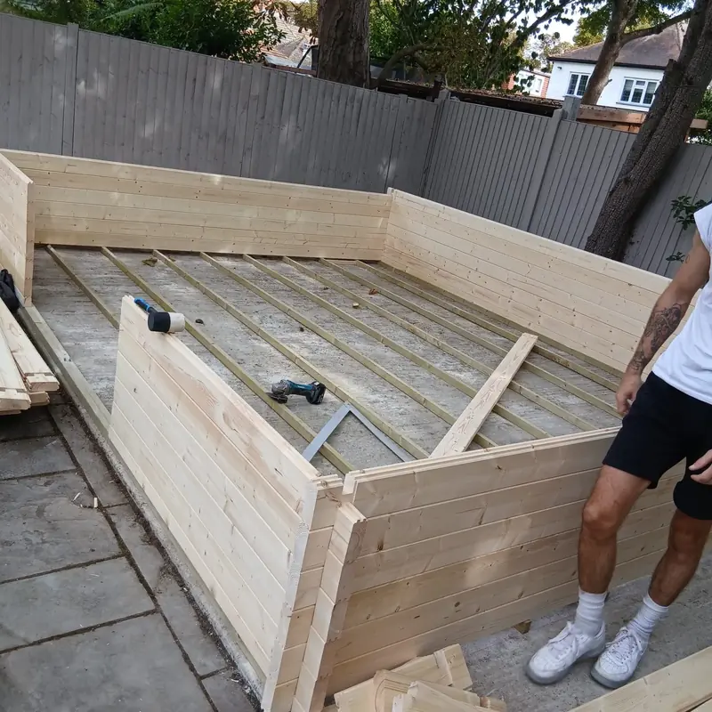 Interlocking log walls rising around timber floor bearers on a concrete base, with an installer beside the front wall.