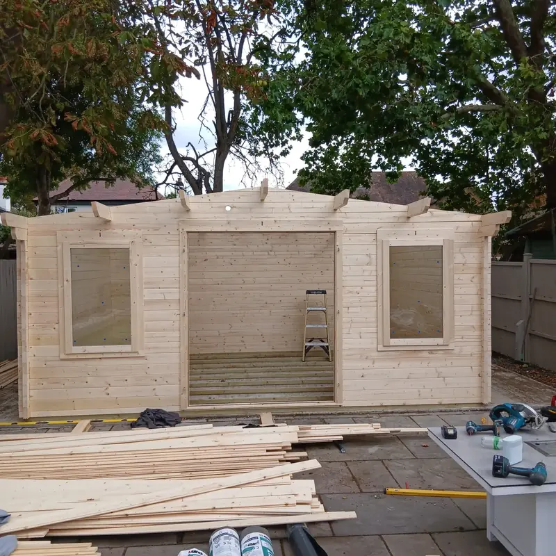 Front of a log cabin being built, with a window either side of a wide double door opening and roof purlins fitted on top