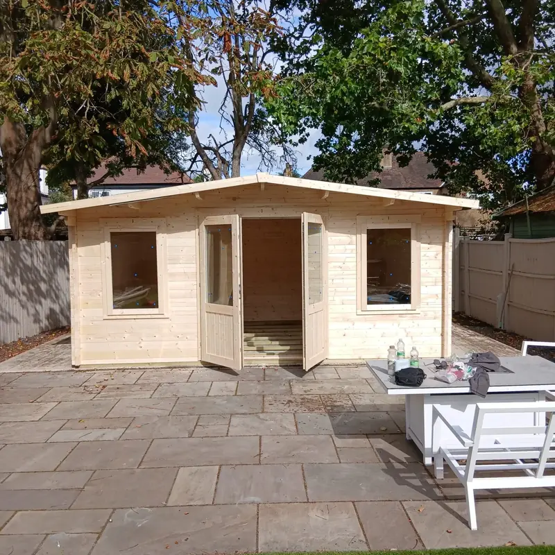 Newly assembled apex log cabin with double doors open and a window either side, on a paved patio under large trees
